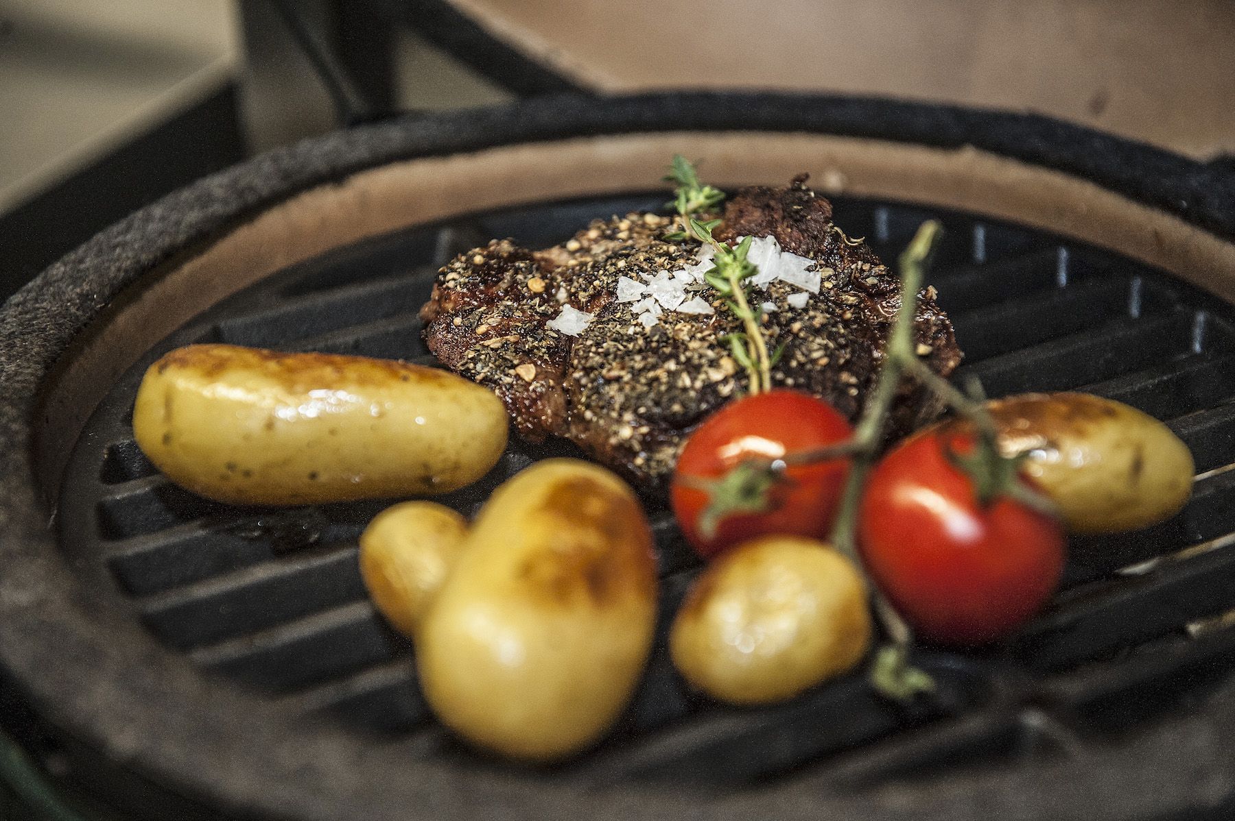 Unterschiedlich grosse Bratkartoffeln zusammen mit einem kleinen Strauch Cherrytomaten und einem Stück gesalzenen und grilliertem Fleisch schmoren auf einer Grillpfanne.